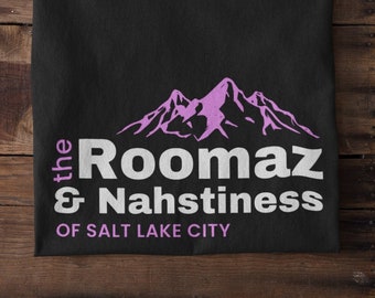 May include: Black t-shirt with the text "the Roomaz & Nahstiness of Salt Lake City" in white and purple. The design includes a purple mountain range graphic above the text. The shirt is folded on a wooden surface.