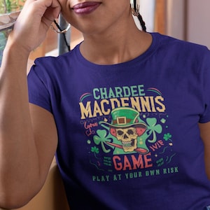 May include: A purple t-shirt with a green, white, and black graphic of a skull wearing a leprechaun hat. The text on the shirt reads "Chardee Macdennis Game Play at Your Own Risk".