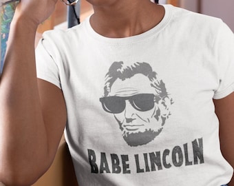 May include: White t-shirt with a gray image of Abraham Lincoln wearing sunglasses and the text "BABE LINCOLN".