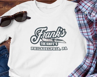 May include: White t-shirt with a vintage-style logo for "Frank's Toe Knife's" in Philadelphia, PA.