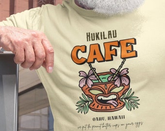May include: A light beige t-shirt with a graphic of a tiki mug with flowers and the text "Hukilau Cafe, Oahu, Hawaii, we put the peanut butter cups on your eggs".