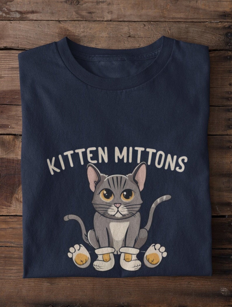 May include: A navy blue t-shirt featuring a cartoon grey cat wearing white booties. The text "Kitten Mittons" is printed on the shirt.
