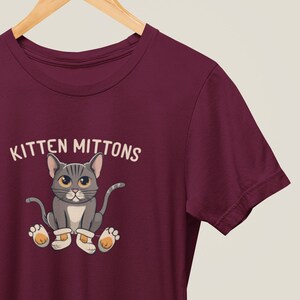 May include: Burgundy t-shirt featuring a cartoon grey cat wearing white mittens and the text "Kitten Mittons".