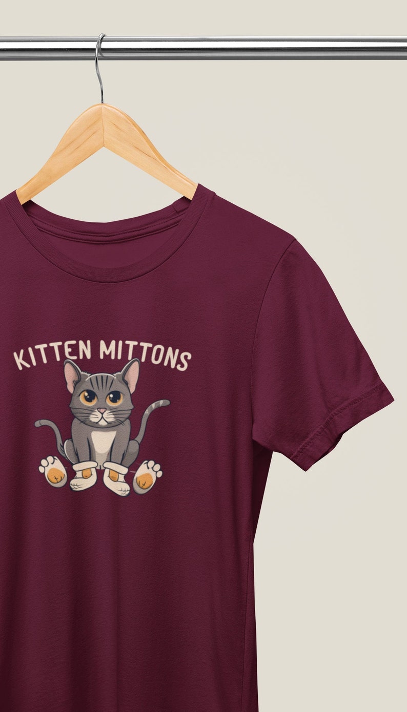May include: Burgundy t-shirt featuring a cartoon grey cat wearing white mittens and the text "Kitten Mittons".