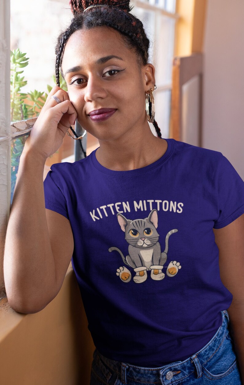 May include: A woman wearing a purple t-shirt with a grey cat wearing white mittens and the text "Kitten Mittons".