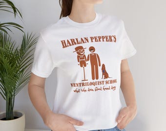 May include: White t-shirt with a brown graphic featuring the text "HARLAN PEPPER'S VENTRILOQUIST SCHOOL" and a stick figure design of a ventriloquist and a dog. The shirt also has the text "what 'chu doin blood hound dogg".