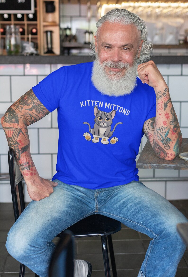 May include: A man wearing a royal blue t-shirt with a cartoon cat wearing mittens and the text "Kitten Mittons".