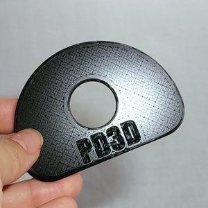 May include: A black plastic half-circle with a hole in the centre and the text "PO3D" printed on the bottom. The surface of the object has a textured pattern.