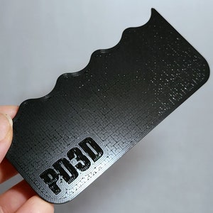 May include: A black, rectangular 3D-printed object with a wavy edge. The text "PD3D" is printed in a bold, blocky font near the bottom. The object has a textured surface and appears to be made of plastic.