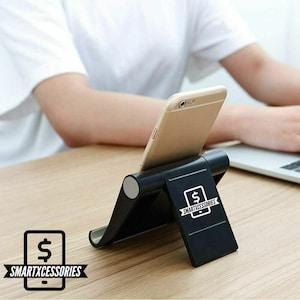 May include: A black foldable phone stand with a silver accent. The stand is holding a smartphone in portrait mode. The stand has a logo that says "SMARTXACCESSORIES" with a dollar sign.