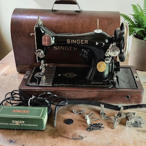 May include: A vintage black Singer sewing machine with gold lettering, housed in a wooden base and carrying case. Includes a green Singer sewing machine attachments box, a black power cord, and several metal presser feet accessories.
