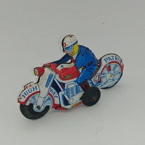 Pode incluir: Uma motocicleta de brinquedo vermelha, branca e azul com uma figura de policial montada nela. A motocicleta tem as palavras "P.D. HIGH WAY" no lado e "PATROL" na roda traseira.