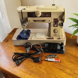 Peut inclure: Une machine à coudre Singer beige dotée d'un panneau de commande marron avec des cadrans et un ensemble d'aiguilles. Sont inclus une pédale noire avec un cordon d'alimentation, un morceau de tissu bleu marine, un tube de lubrifiant et des canettes.