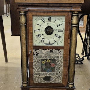 Puede incluir: Reloj antiguo de madera con esfera crema y números romanos negros. El reloj tiene un panel floral decorativo debajo de la esfera y columnas doradas. El reloj mide aproximadamente 40 cm de altura.