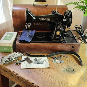Peut inclure: Une machine à coudre Singer noire vintage avec une base en bois et un couvercle arqué assorti. La machine présente des lettres dorées, un cordon d'alimentation et un morceau de tissu bleu sous l'aiguille. Accessoires : boîte verte et manuel.