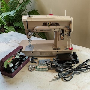 Puede incluir: Una máquina de coser Singer de color beige con pedal negro, cable de alimentación y un estuche de accesorios granate que contiene bobinas de metal y un accesorio para ojales. Incluye una caja con la etiqueta Singer Templates y bobinas.