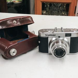 May include: A silver and black Voigtlander camera with a brown leather case. The camera has a lens with a black cap and the text "Voigtlander" printed on the front.