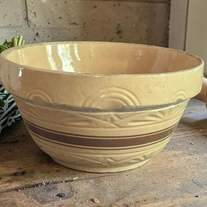 May include: A cream-coloured ceramic mixing bowl featuring a wide rim, a smooth interior, and an exterior decorated with embossed semi-circular arches and a horizontal brown band. The bowl has a thick, sturdy construction with a glossy finish.