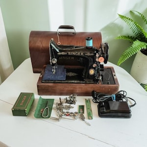 May include: A vintage black Singer sewing machine with gold lettering, housed in a wooden base and cover. Included are a green Singer box, various metal sewing attachments, bobbins, a small tool, and a black electric foot pedal with a power cord.