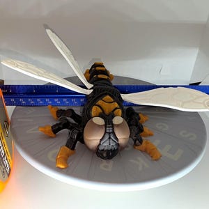 May include: A detailed, plastic insect model with black, yellow, and tan coloring. The wings are white. The model is approximately 4 inches long, as measured by a yellow tape measure.