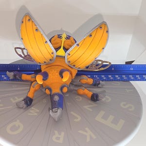 May include: A detailed, orange and blue insect-like figurine with large yellow wings. The figurine is positioned on a gray surface with letters and numbers. A blue ruler is placed behind the figurine.