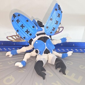 May include: A handmade insect sculpture with blue wings and accents, black and white body, and six legs. The wings have a cross pattern. A blue ruler with inch measurements is next to the sculpture.