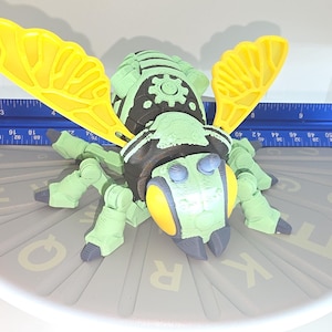 May include: A green and black mechanical bee toy with yellow wings. The toy is on a gray surface with letters and numbers. A blue ruler is next to the bee. The bee measures approximately 6 inches long.