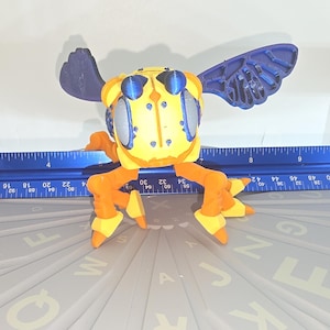 May include: A yellow and orange toy insect with blue wings and large blue eyes. The toy is positioned on a gray surface with a blue ruler. The ruler measures up to 25 inches.
