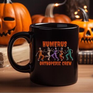 K&ouml;nnte beinhalten: Schwarze Keramik-Tasse mit dem Text "HUMERUS ORTHOPEDIC CREW" und einer Grafik von tanzenden Skeletten in Orange, Lila und Gr&uuml;n. Der Hintergrund zeigt geschnitzte orangefarbene K&uuml;rbisse, was auf ein Halloween-Thema hindeutet.
