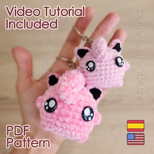 Könnte beinhalten: Zwei rosa gehäkelte Schlüsselanhänger in Form von Zeichentrickfiguren, jeweils mit schwarzen Akzenten und großen, ausdrucksstarken Augen. Die Schlüsselanhänger werden von einem silbernen Schlüsselring gehalten. Der Text "Video Tutorial Included" und "PDF Pattern" ist enthalten.
