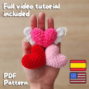 May include: Three crocheted hearts, one pink with white wings, one pink, and one red. The hearts are held in a hand. The text "Full video tutorial included" and "PDF Pattern" are at the top and bottom of the image. The image also includes flags for the United States and Spain.