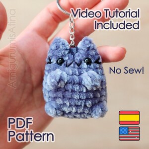 Könnte beinhalten: Ein blauer, plüschiger, gehäkelter Katzen-Schlüsselanhänger mit schwarzen Perlenaugen und gestickten Schnurrhaaren. Das Bild enthält den Text "Video Tutorial Included", "No Sew!", "PDF Pattern", "AmigurumisAtina" sowie spanische und amerikanische Flaggen.