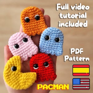 Puede incluir: Personajes de Pac-Man y fantasmas de crochet en varios colores, incluyendo amarillo, naranja, rosa, azul y rojo. La imagen incluye el texto "Tutorial de video completo incluido" y "Patrón PDF". También se incluyen las banderas de España y los Estados Unidos.