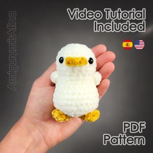 May include: A handmade white crochet duck with a yellow beak and feet. The duck is being held in a hand. Text on the image says "Video Tutorial Included" and "PDF Pattern".