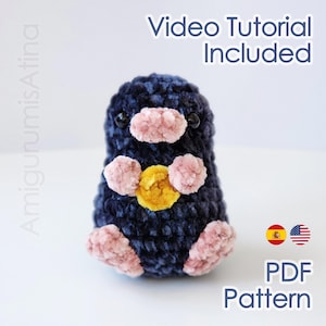 Puede incluir: Una criatura amigurumi de color azul oscuro con detalles rosas en la nariz, las patas y el vientre. Tiene ojos negros y un círculo amarillo en el pecho. La imagen incluye el texto "Video Tutorial Included" y "PDF Pattern".