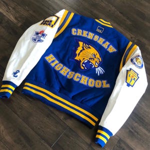 Può includere: Giacca college blu e bianca con dettagli gialli. Il retro presenta "CRENSHAW HIGH SCHOOL" e un'immagine di tigre. Toppe sulle maniche con testo e loghi. La giacca ha colletto, polsini e orlo a coste.