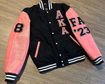 Op de afbeelding: Zwarte en roze varsity jas met de letters "AKA" en "FA 23" op de mouwen. De jas heeft een zwarte body, roze mouwen en wit-zwarte gestreepte manchetten en kraag. Het nummer "8" staat op de linkermouw.