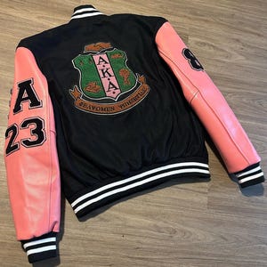 Op de afbeelding: Zwarte varsity jas met roze mouwen. Voorzien van de letters "A" en "23" op de linkermouw en het nummer "8" op de rechtermouw. De achterkant toont een wapen met de letters "AKA" en Griekse tekst. Wit en zwart gestreepte bies.