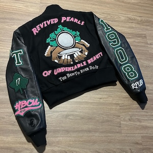 May include: Black varsity jacket with leather sleeves. The back features the text "REVIVED PEARLS" and "OF UNDENIABLE BEAUTY" in pink, along with a graphic. The left sleeve has a "T" and "17" patch. The right sleeve has "1908" in green.