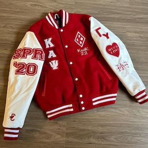 Könnte beinhalten: Eine rot-weiße Collegejacke mit mehreren Aufnähern und Text. Die Jacke hat weiße Ärmel mit rot-weiß gestreiften Bündchen. Zu den Aufnähern gehören "SPRAY 20", "K A Ψ", eine Raute und ein Herz mit "MUCH LOVE".