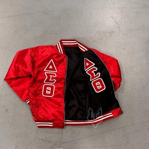 Op de afbeelding: Een rode satijnen jas met wit en rood gestreepte bies. De jas heeft de Griekse letters Delta, Sigma en Omega in het wit op de voorkant. De binnenkant van de jas is zwart.