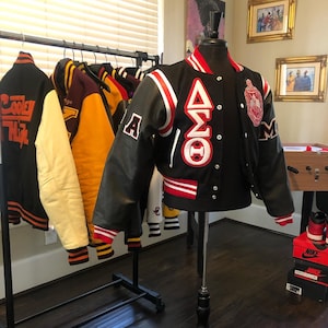 May include: Black varsity jacket with white and red detailing, displayed on a mannequin. The jacket has Greek letters and patches. Other varsity jackets in various colours hang on a rack in the background.