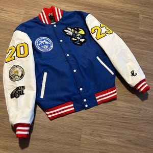 May include: A blue varsity jacket with white sleeves, featuring patches and the numbers "20" and "23" in yellow. The jacket has red and white striped trim on the collar, cuffs, and hem. Patches include a bee mascot and a school emblem.