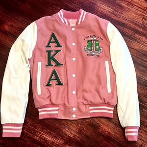 Op de afbeelding: Roze en witte varsity jas met de letters AKA in groen op de voorkant. De jas heeft witte mouwen en een roze body. De jas is ook voorzien van het Alpha Kappa Alpha Sorority, Inc. embleem.