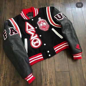 May include: Black varsity jacket with red and white detailing. It has leather sleeves, a red and white striped collar and cuffs, and patches with Greek letters and a crest. The jacket fastens with buttons.