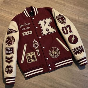 Puede incluir: Una chaqueta universitaria granate con mangas color crema, que presenta una letra K grande, una antorcha y un parche de corona. La chaqueta incluye textos como Your Name, EST. 2024, CAPTAIN, TEAM WORK, MVP, ATHLETICS, 07, LEGACY y CLASS OF 24 con un balón.