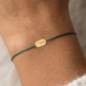 Pode incluir: Uma pulseira dourada com o nome "Laura" gravado em um pequeno pingente oval. O pingente está preso a um cordão verde escuro. A pulseira é usada no pulso, com um suéter branco ao fundo.