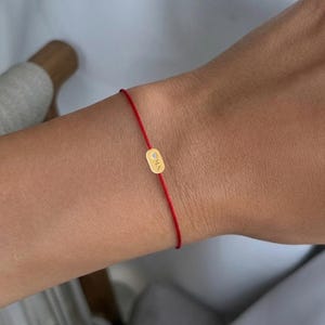 Puede incluir: Una pulsera de hilo rojo con un pequeño dije rectangular de color dorado. El dije tiene las letras "ONA" grabadas. La pulsera se lleva en la muñeca.