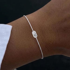 Könnte beinhalten: Ein zartes Armband aus weißem Kordel mit einem kleinen, silberfarbenen Anhänger. Der Anhänger hat ein dezentes Design, das dem minimalistischen Accessoire einen Hauch von Eleganz verleiht. Das Armband wird am Handgelenk getragen, was auf Schmuck und persönlichen Schmuck hindeutet.