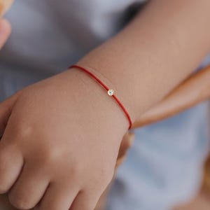 Può includere: Un delicato braccialetto con cordino rosso caratterizzato da una singola piccola pietra preziosa rotonda e trasparente incastonata in una montatura color oro. Il filo sottile avvolge il polso, offrendo un look essenziale con un punto luce.